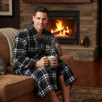 A man wearing a plush black and white plaid bathrobe sits on a brown leather couch in a cozy living room, holding a mug with steam rising. A stone fireplace with a burning fire is visible in the background.