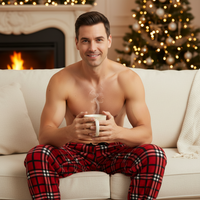 A shirtless man sitting on a white couch in front of a fireplace, holding a steaming mug. He is wearing red plaid pajama pants, with a decorated Christmas tree and warm holiday lights in the background.
