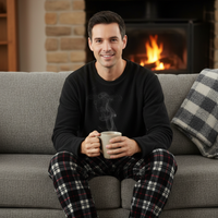 A man wearing a black long-sleeve pajama top and red/black/white plaid pajama pants sits barefoot on a gray couch in front of a lit fireplace, holding a mug.