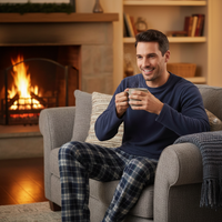 A man sitting in a cozy gray armchair by a lit fireplace, wearing a navy long-sleeve shirt and blue plaid pajama pants while holding a mug. The background shows a warm, decorated living room with bookshelves and a glowing fire.