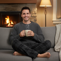 A man sitting cross-legged on a gray couch in front of a lit fireplace, wearing a dark gray long-sleeve shirt and dark plaid pajama pants while holding a light beige mug with steam rising.
