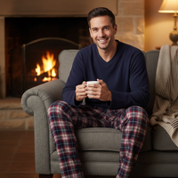 A smiling man sits on a gray armchair in front of a lit fireplace, holding a white mug. He wears a navy blue long-sleeve shirt and red-and-blue plaid pajama pants, with a cream throw blanket draped over the chair.