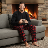 A man sits on a gray sofa in front of a lit fireplace, wearing a black long-sleeve shirt and red plaid pajama pants while holding a mug.