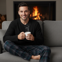 A man sitting cross-legged on a gray couch in front of a lit fireplace, wearing a black long-sleeve shirt and dark plaid pajama pants while holding a white mug.