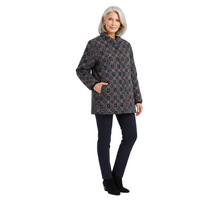 A woman modeling a dark patterned jacket with a stand collar, paired with navy pants and black ankle boots, against a plain light gray studio background.