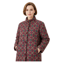 A woman wearing a high-neck, button-front coat with a dense floral and medallion pattern in burgundy, green, and beige tones, photographed against a plain light gray studio background.