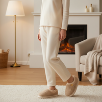 A person wearing a cream-colored two-piece pajama set and matching fuzzy slip-on slippers stands in a cozy living room with a fireplace, lamp, and armchair.