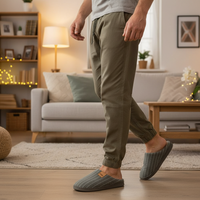 A man wearing olive green jogger-style pants and gray slip-on slippers with thin white pinstripes, standing in a cozy living room with a sofa, lamp, and wooden furniture in the background.