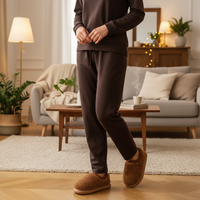 A person wearing a matching dark brown lounge set and fuzzy brown slip-on slippers stands in a cozy living room with a sofa, lamp, coffee table, and plants in the background.
