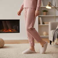 A person wearing a matching dusty pink lounge set with ribbed knit pants and a long-sleeve top, paired with light beige slip-on slippers, standing in a cozy living room with a fireplace and lamp in the background.