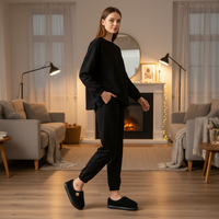 A woman stands in a warmly lit living room wearing a black long-sleeve top and black jogger-style pants, with black slip-on slippers. The room has gray sofas, curtains, a fireplace, and floor lamps.