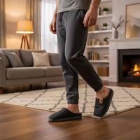 A person wearing dark gray jogger-style slip-on slippers and a matching gray top stands in a cozy living room with wood floors, a rug, a sofa, shelves, and a fireplace.