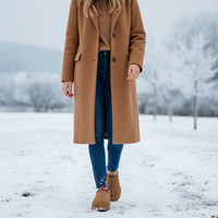 A person standing outdoors in a snowy landscape wearing a long camel-brown coat over a tan sweater, blue jeans, and tan boots.