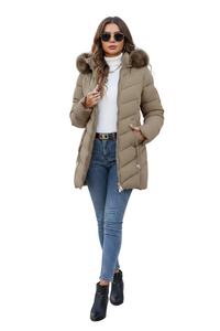 A woman modeling a mid-length beige puffer jacket with a brown faux-fur trimmed hood, worn over a white turtleneck and blue jeans, on a solid white background.