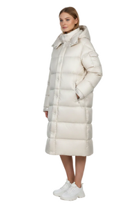 A woman modeling a long, puffer-style hooded coat in a warm off-white or cream color, paired with white sneakers, against a plain white studio background.