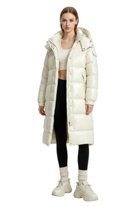 A model stands against a light gray studio background wearing a long, glossy puffer coat in off-white/cream. The coat is quilted with a large hood and front snap buttons.