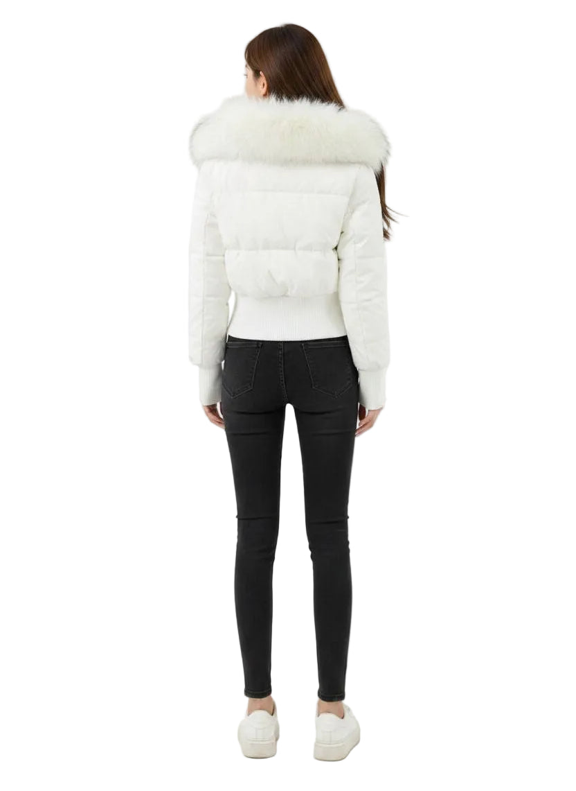 A woman viewed from the back wearing a white puffer jacket with a large fluffy faux-fur collar, black skinny jeans, and white sneakers, set against a plain white studio background.