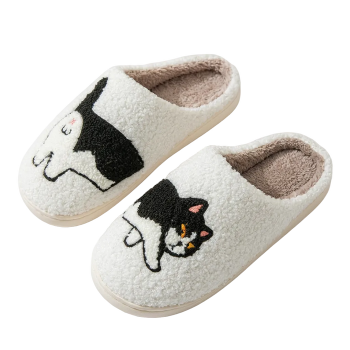 A pair of fluffy slip-on house slippers in off-white sherpa, each decorated with a black-and-white cat illustration and a soft tan lining, photographed on a clean white background.