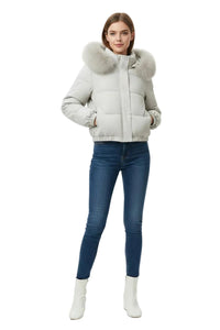 A woman modeling a light gray puffer jacket with a large fluffy faux-fur collar, paired with dark blue skinny jeans and white ankle boots, against a plain white studio background.