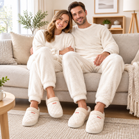 A smiling couple wearing matching cozy cream fleece pajama sets and quilted slip-on slippers sits together on a light beige sofa in a bright, modern living room.
