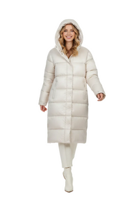 A woman modeling a long, hooded puffer coat in a warm off-white or cream color, paired with matching cream pants and white ankle boots, against a seamless white studio background.