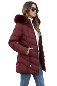 A woman wearing a long quilted puffer coat in burgundy with a faux-fur trimmed hood, styled with a white turtleneck, sunglasses, and blue jeans against a plain white background.