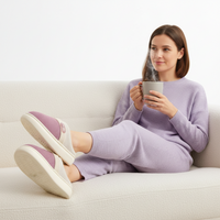 A woman sits relaxed on a light beige sofa holding a gray mug, wearing a matching lavender waffle-knit lounge set and mauve slip-on slippers with cream soles.