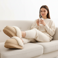 A woman sits on a light beige sofa wearing chunky beige slip-on slippers with white faux-fur trim and a matching cream knit sweater and pants, holding a white mug.