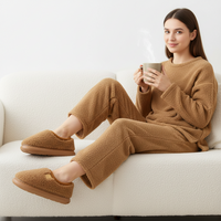 A woman sits on a white textured sofa wearing a matching tan fleece pajama set and slip-on slippers, holding a steaming mug.