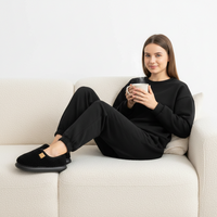 A woman sits on a cream-colored textured couch, wearing a matching black sweatshirt and jogger pants with black slip-on slippers. She is holding a white mug.
