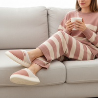 A woman sits on a light gray sofa wearing a matching pink and white striped knit sweater and slip-on slipper set, holding a white mug.