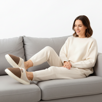A woman sits on a light gray sofa wearing a matching cream sweatshirt and jogger pants with brown slip-on shoes. The setting is a clean, minimalist studio with a white wall background.