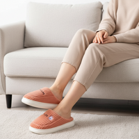 A person seated on a light beige sofa wearing ribbed slip-on house slippers with white soles. The slippers are a muted coral pink color with a small embroidered character detail on the front.