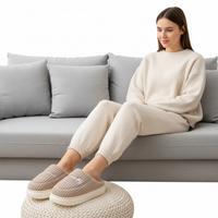 A woman sits on a light gray sofa wearing a cream ribbed knit sweater and matching jogger pants, with beige slip-on slippers on a woven ottoman. The background is a clean white studio setting.