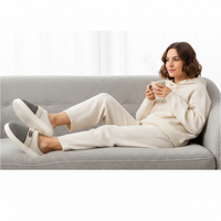 A woman reclining on a light gray textured couch, wearing a matching cream-colored lounge set and slippers while holding a white mug.