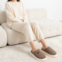 A person sitting on a cream tufted sofa wearing a matching cream knit lounge set and brown slip-on slippers with white soles, in a bright indoor setting with a white rug.