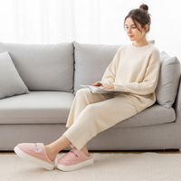 A woman sits on a light gray sofa reading a magazine, wearing a cream-colored knit lounge set and pink slip-on slippers with white soles in a bright, modern living room.