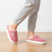 A person wearing pink ribbed slip-on house slippers with white soles and a small bunny patch on one slipper, shown standing on a light wood floor against a white wall.