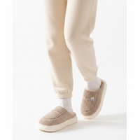 A person wearing beige ribbed slip-on slippers with white socks and cream jogger pants, shown from the calves down against a clean white studio background.