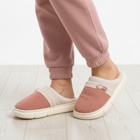 A person wearing pink knit slip-on mule slippers with cream elastic bands and cream soles, paired with matching pink jogger pants, standing on a light wood floor.