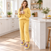 A woman stands in a bright, modern kitchen wearing a fuzzy yellow two-piece pajama set with matching slip-on socks. She holds a white mug, and the kitchen features white cabinets, a marble island, and warm sunlight streaming in from a window.